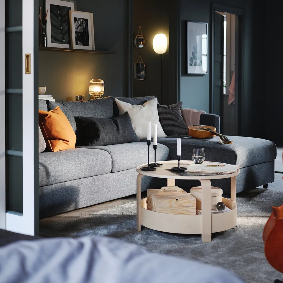 Modern living room with grey VIMLE sofa, orange throw pillows, wooden coffee table, candles, and dark walls.