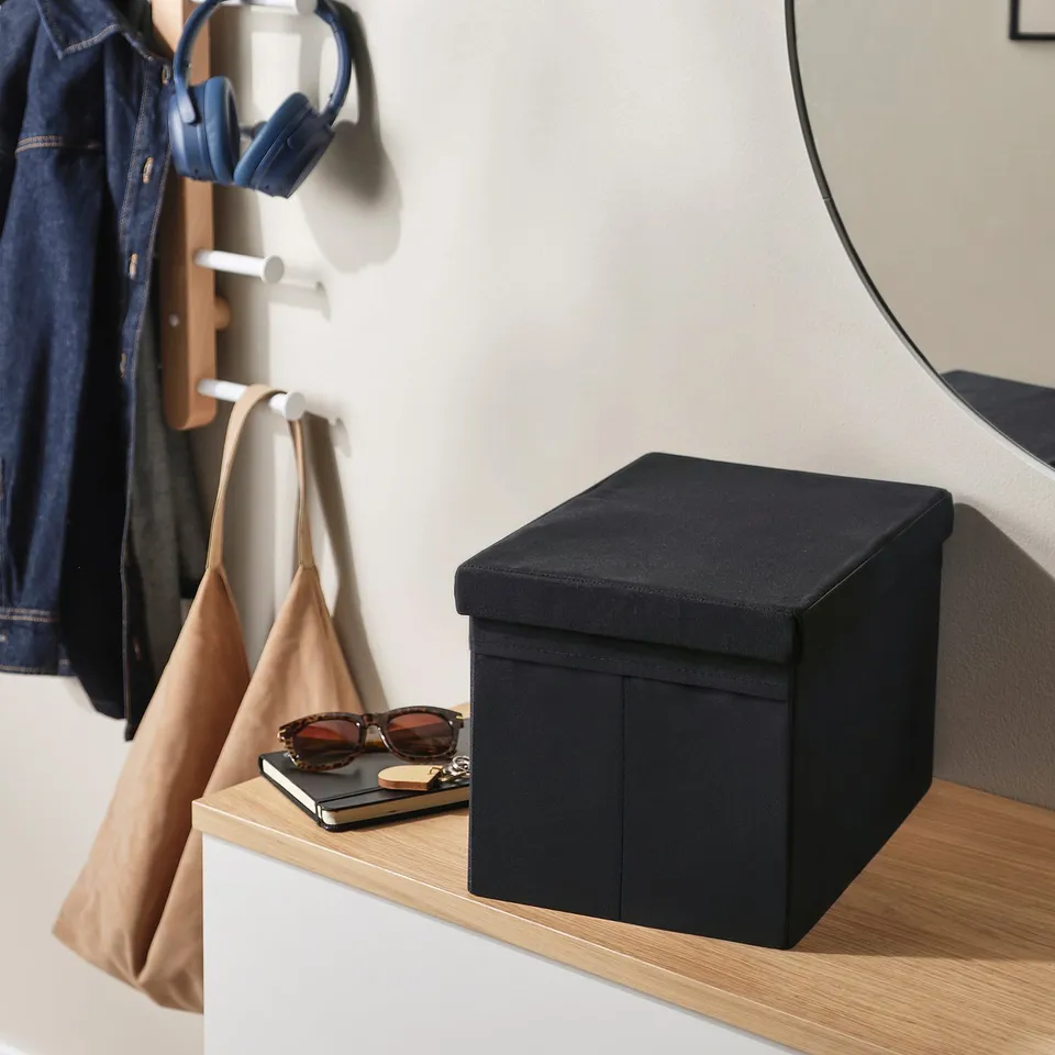 Black fabric storage box on shelf, with sunglasses and book on top. Hanging coat and shoulder bag nearby.