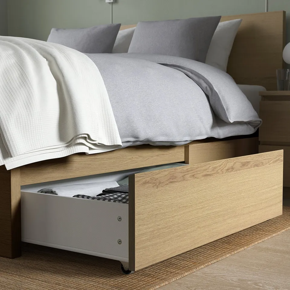Bed with open drawer under frame, showing folded clothes; light wood finish, grey bedding.