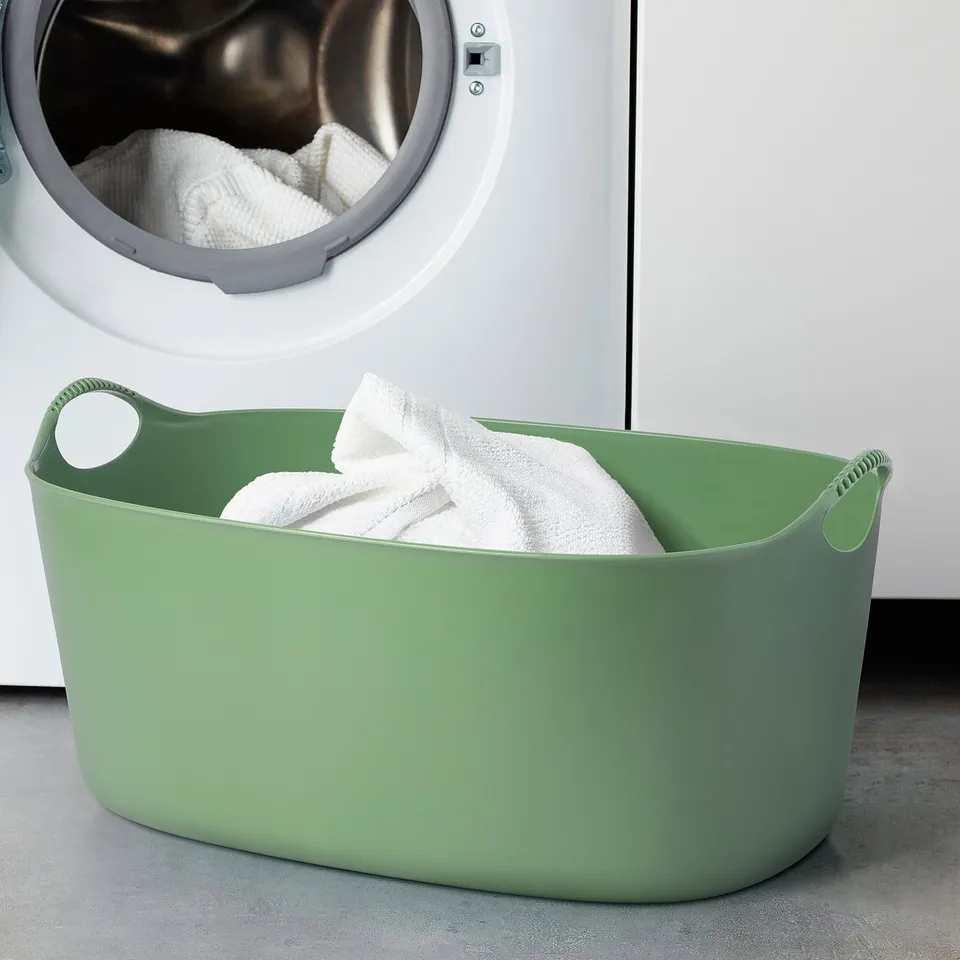 A green TORKIS laundry basket holding towels, positioned in front of a washing machine.