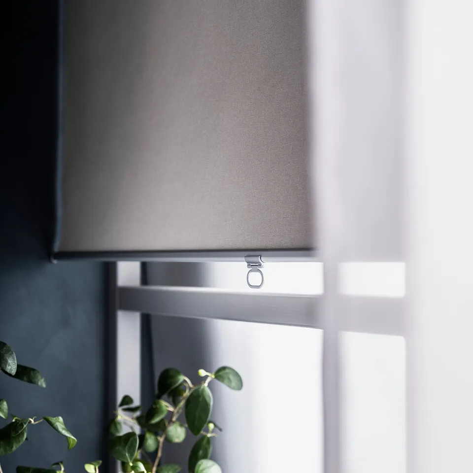 A cordless grey roller blind, SKOGSKLÖVER, is partially open with a minimalistic design, mounted inside a window. An indoor plant is visible below.