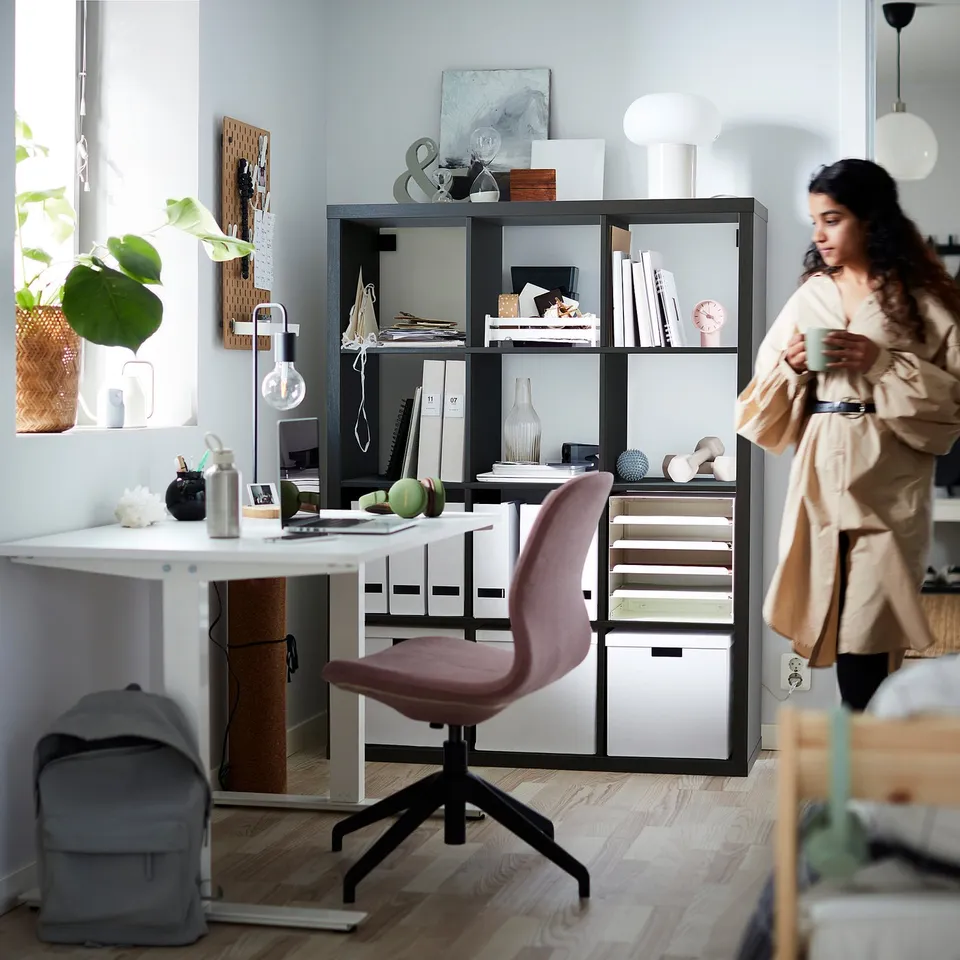 Person at tidy home office with KALLAX shelf, desk, chair, laptop, lamp, backpack, and plant.