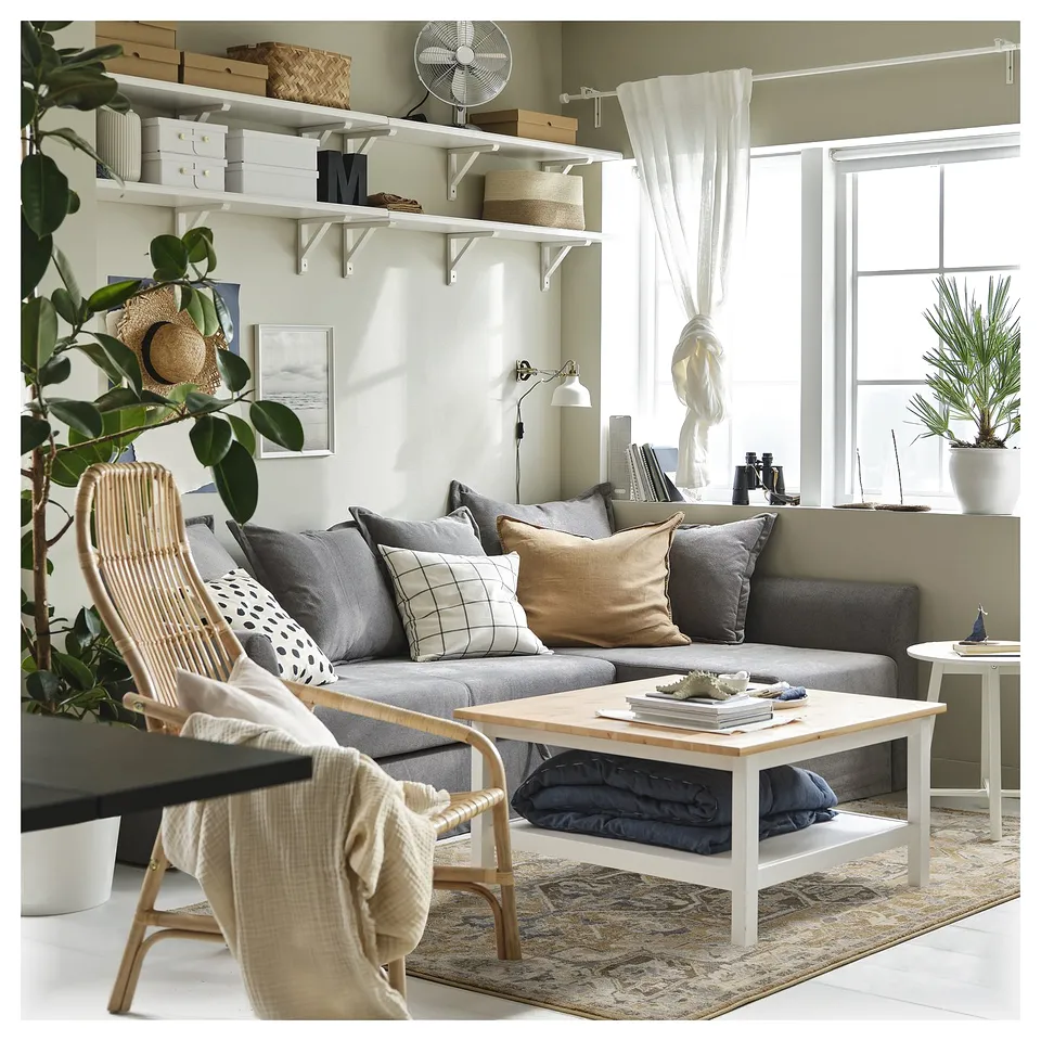Depicts a cosy living room with pastel-colored rugs, grey couch, wooden coffee table, wicker chair, and shelves with decor.