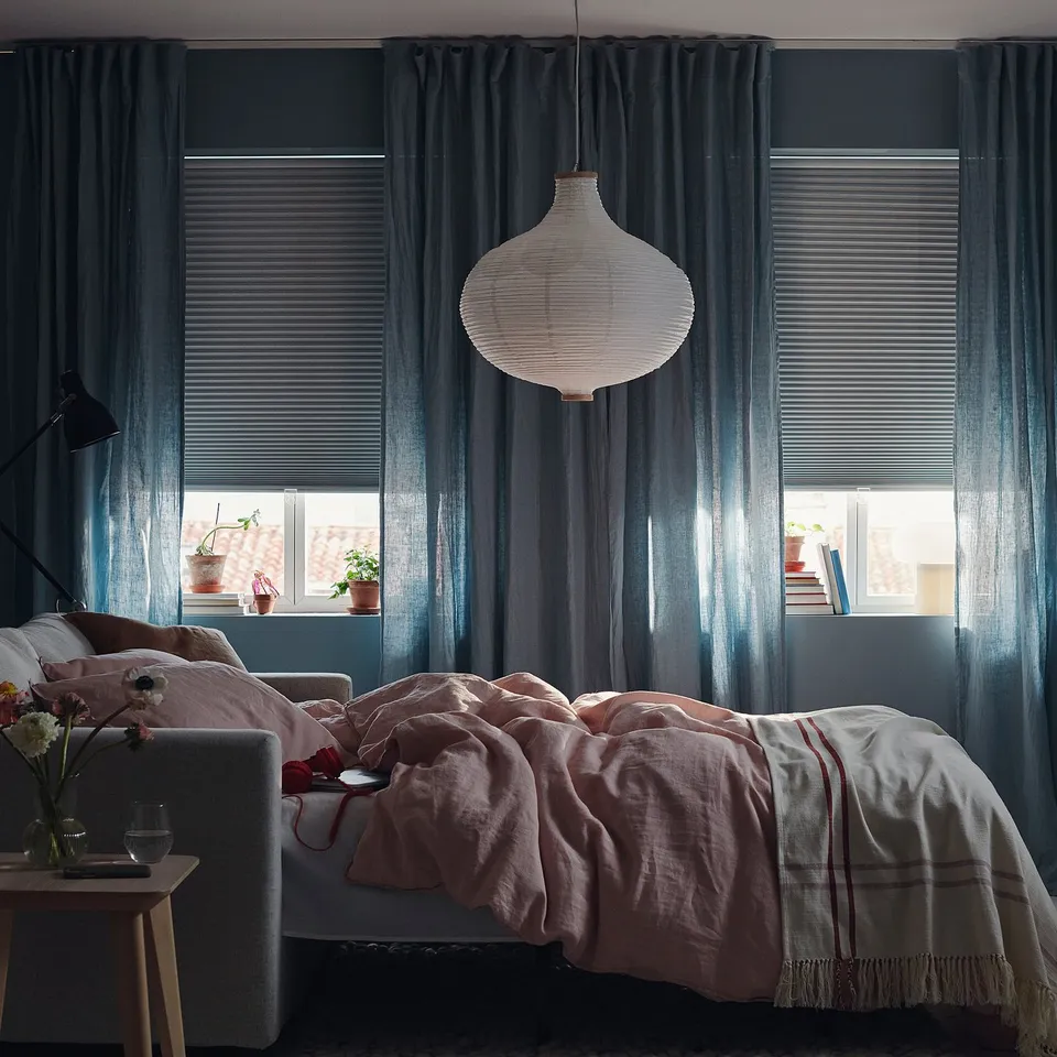 Bedroom with large windows, wooden blinds, and hanging light. Curtains block light, and bed has pink blankets.