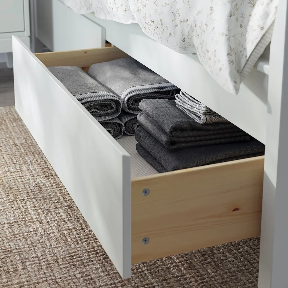 Open white wooden drawer, neatly folded grey towels, under a bed with a white floral pillow.