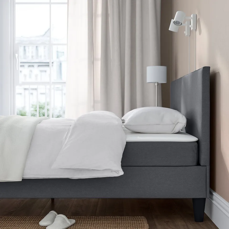 Bed with grey upholstered headboard, white linens, wall lamp, and slippers on a mat, situated by a large window.