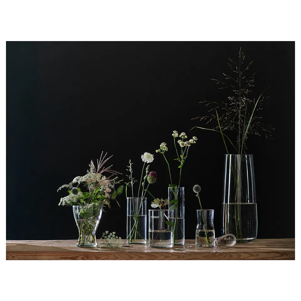 Clear glass BERÄKNA vases in various shapes and sizes, holding delicate white flowers and dried grasses, arranged on wooden table against dark wall.