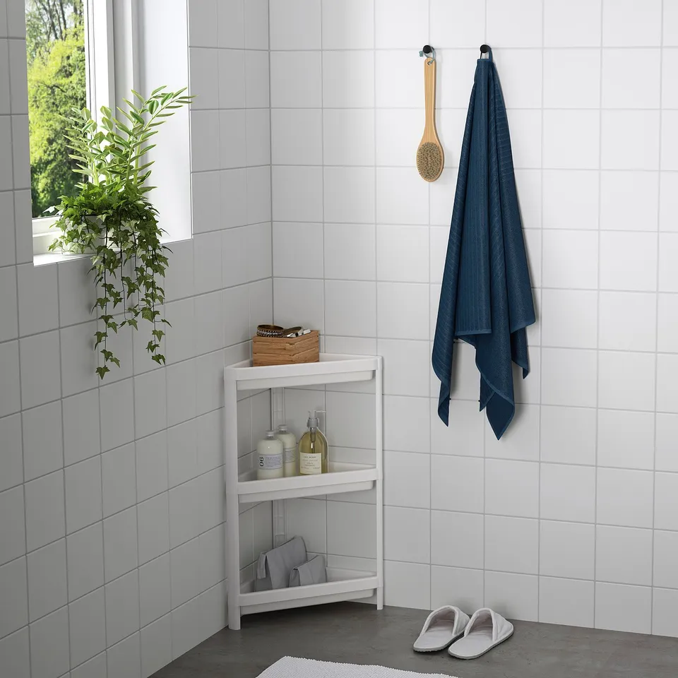 A small, white, three-shelf unit in a bathroom with toiletries, a plant, and a towel hanging nearby.