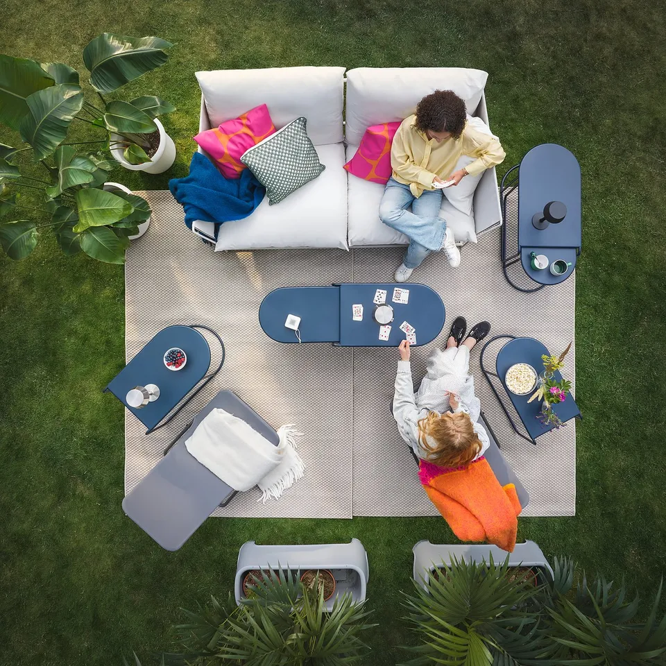 Two playing cards outdoors on rug with cushions. One in chair, one on rug. Cups, folded chair, plants.