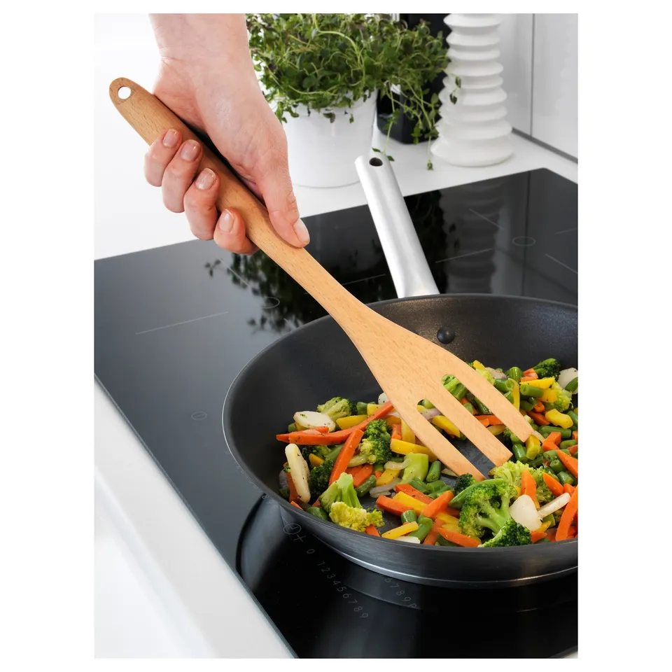 A hand holding a wooden RÖRT spatula stirs colourful vegetables in a black pan on a stove. The spatula is kind to non-stick surfaces.