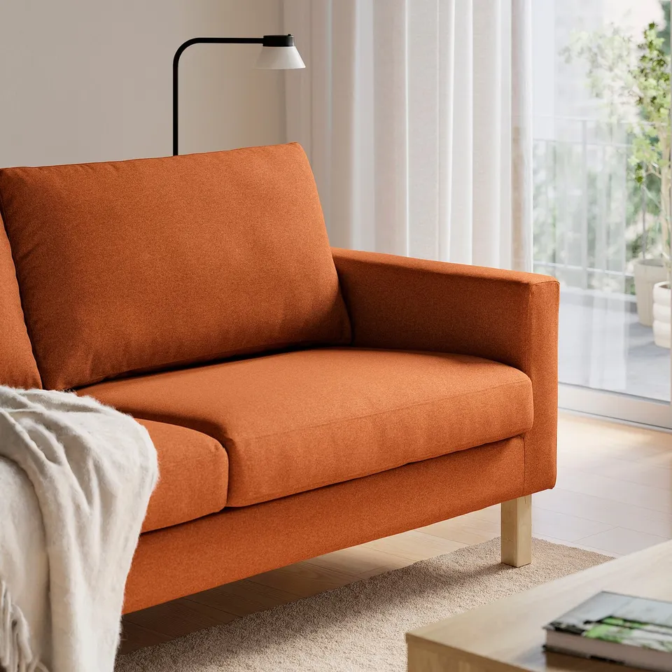 Orange upholstered loveseat with wooden legs next to white lamp and window.