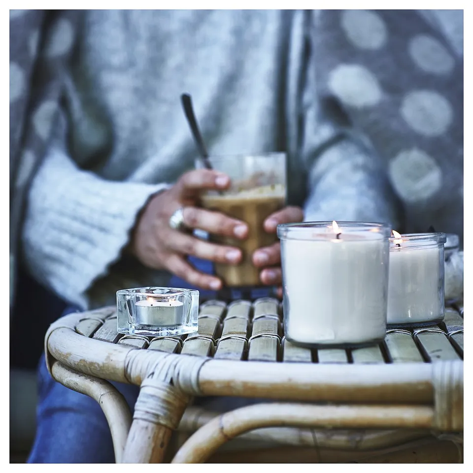 A person sits near lit candles on a wicker table. One candle is in a glass, others in clear holders. Soft light casts a warm glow.