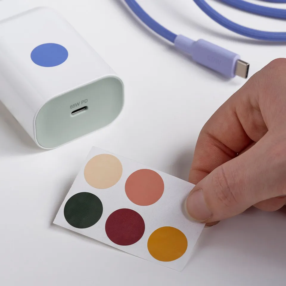A hand holds a sheet of coloured stickers beside a 65w usb-c charger and a purple charging cable.