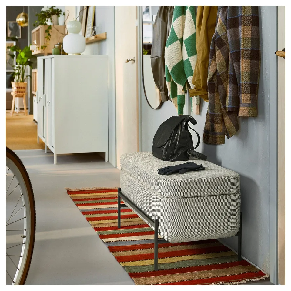 Cosy room with VALTORP bench, striped rug, and plaid jackets. Bag & gloves on bench, cabinet with decor, bike wheel.