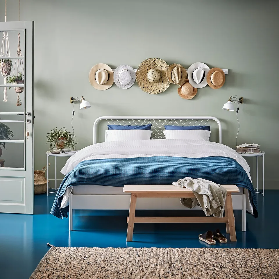 Modern bedroom with white metal bed, blue bedding, wall-mounted hats, plants, and blue floor.