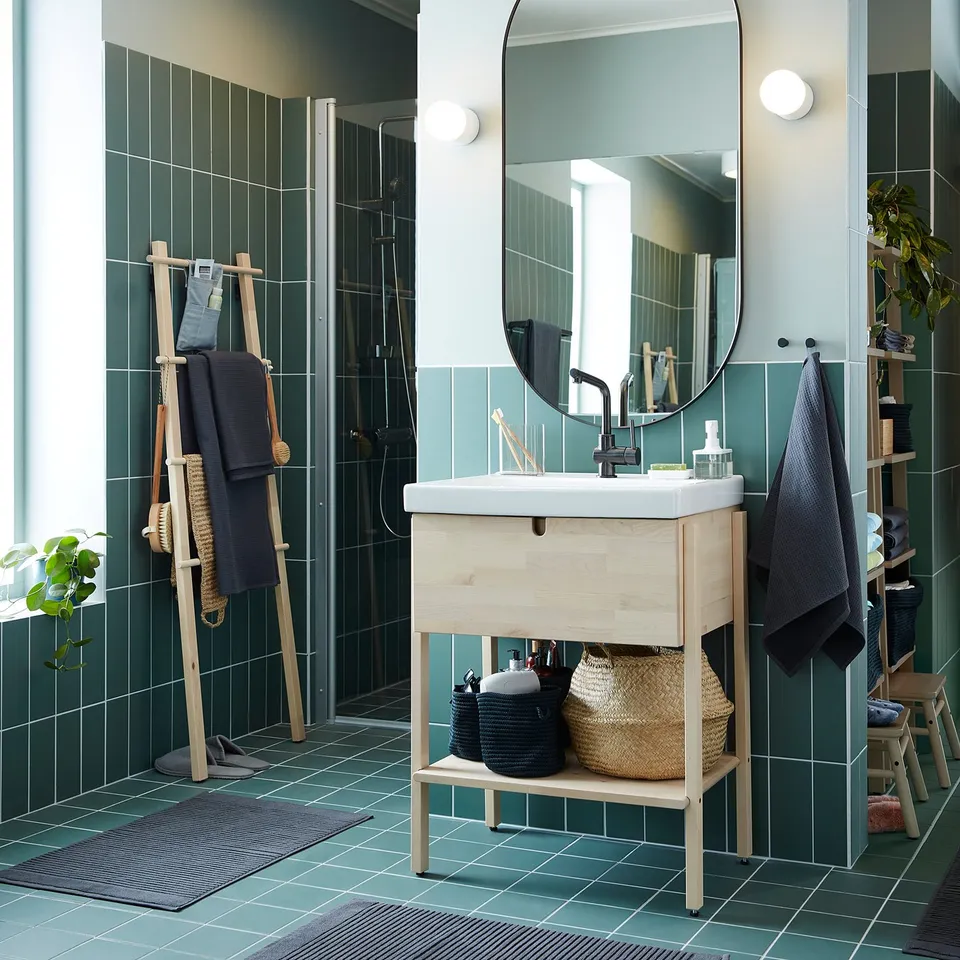 Modern bathroom with light wood vanity, green tiles, black towels, and a large round mirror with a black frame.