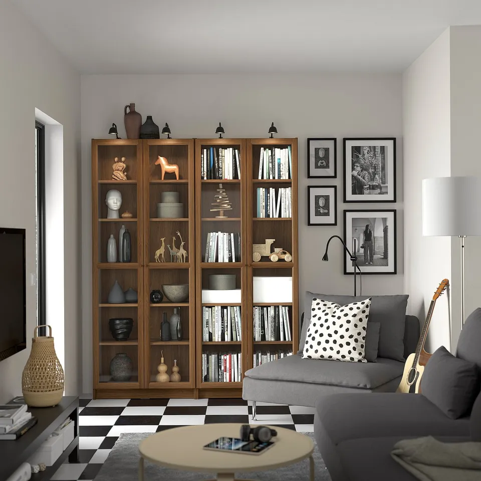 BILLY bookshelf with glass, books & decor next to grey sofa with spotted pillow. Chequered floor, black & white wall photos.