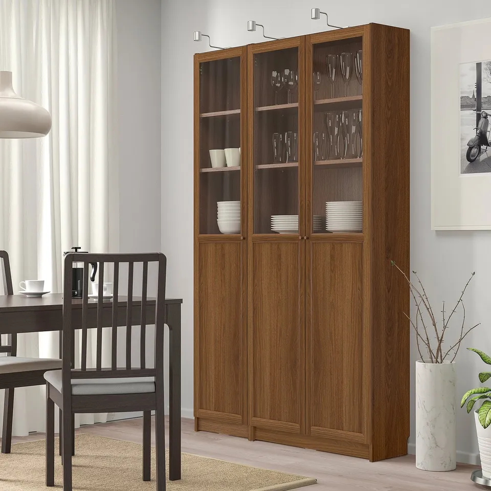 Large BILLY wooden bookcase with glass doors and brown stain. Tall, holding glasses and plates, beside black dining chairs.