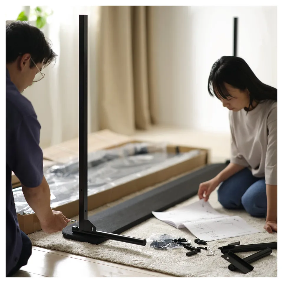 Two people assemble a flat-packed SLATTUM bed frame; dark grey, modern design with upholstered headboard and steel slats.