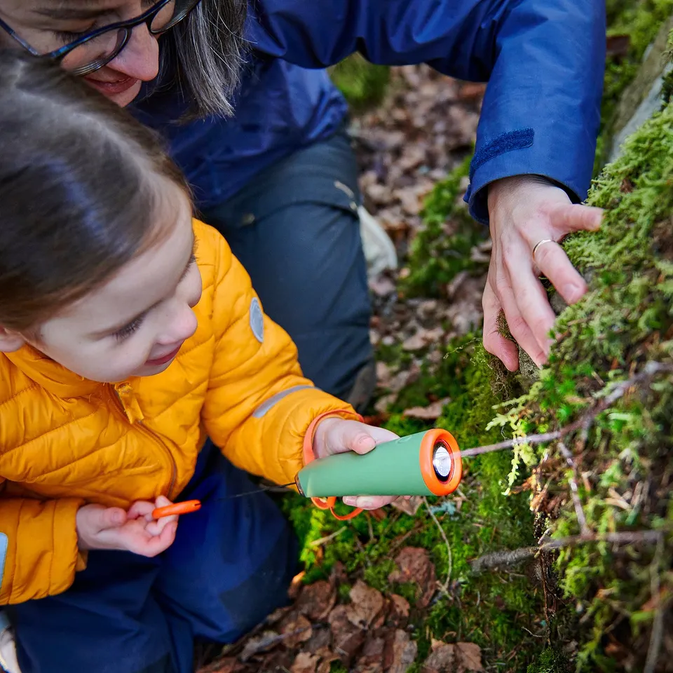 Child and adult examine moss with green torch, highlighting outdoor exploration.