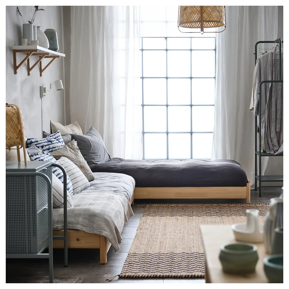 Modern bedroom with two wooden UTÅKER beds pushed together, styled with pillows and a rug. Large window with curtains.
