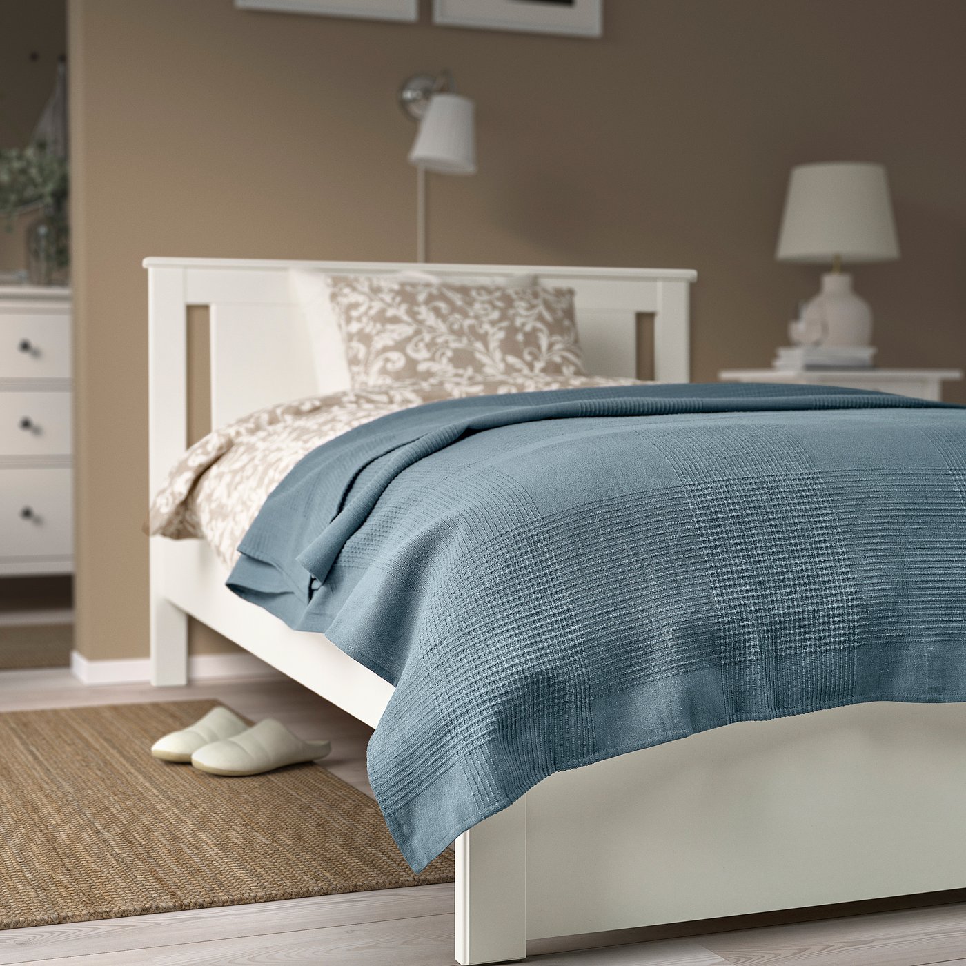 Blue cotton bedspread on large white bed, with pillows, lamp, and slippers nearby.