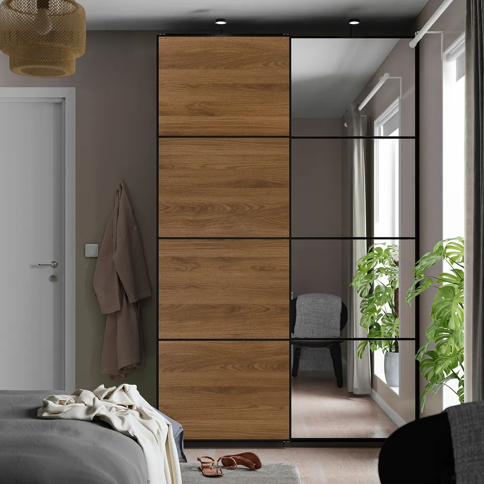 Modern bedroom with large wooden and mirror sliding wardrobe, grey bed, and a large plant. Wardrobe is taller than bed, with clothes on hooks.