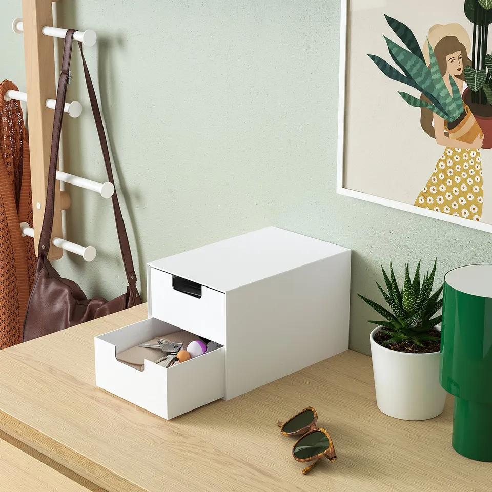 White mini storage chest on desk with open drawer showing small items, next to plant and artwork.