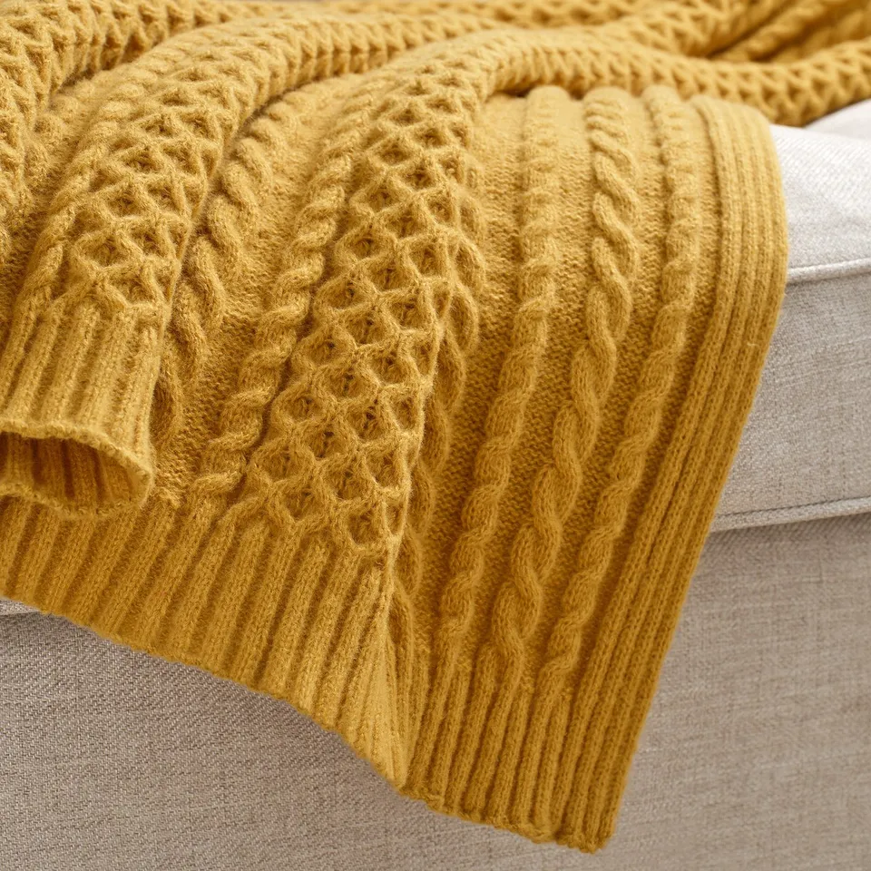 Golden mustard yellow knitted blanket with intricate patterns draped cosily on a beige sofa, showcasing its soft texture and elaborate design.