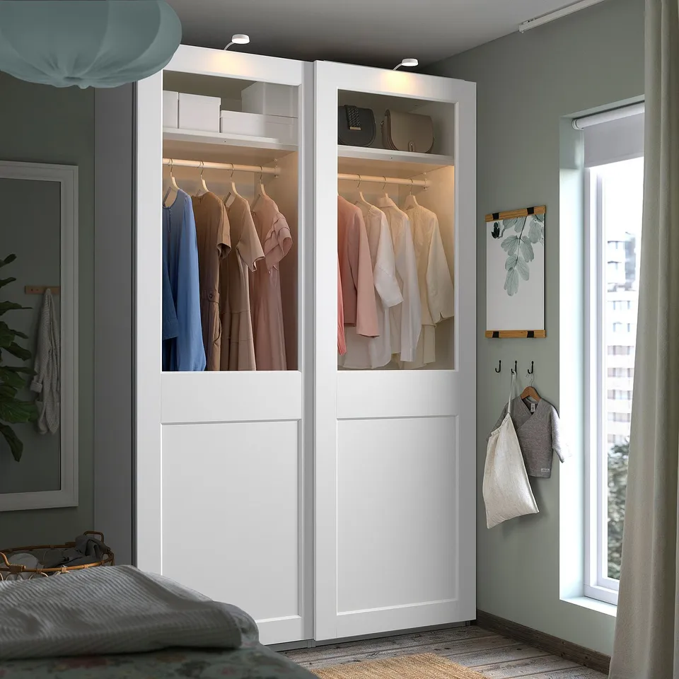 Modern bedroom featuring a large, white PAX wardrobe with glass doors showcasing clothes, situated next to a large window with curtains.