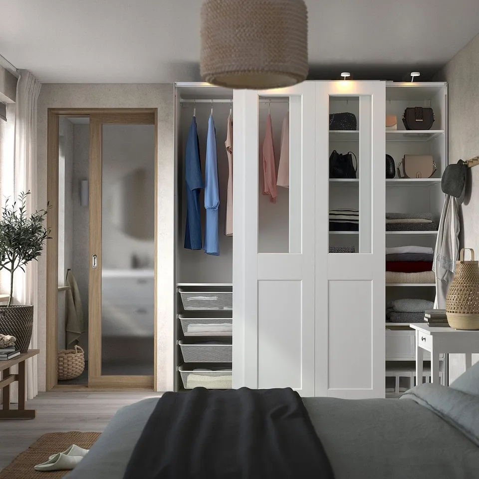 Modern bedroom with white PAX wardrobe, hanging clothes, shelves, and open storage. Black blanket, wicker baskets, and plants create cosy space.
