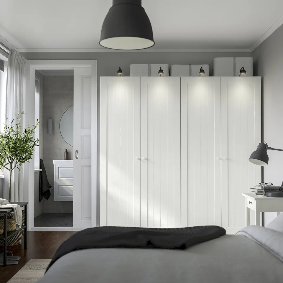 Modern bedroom with large white wardrobe, grey boxes, sleek bathroom vanity, and made bed with black and grey linens.