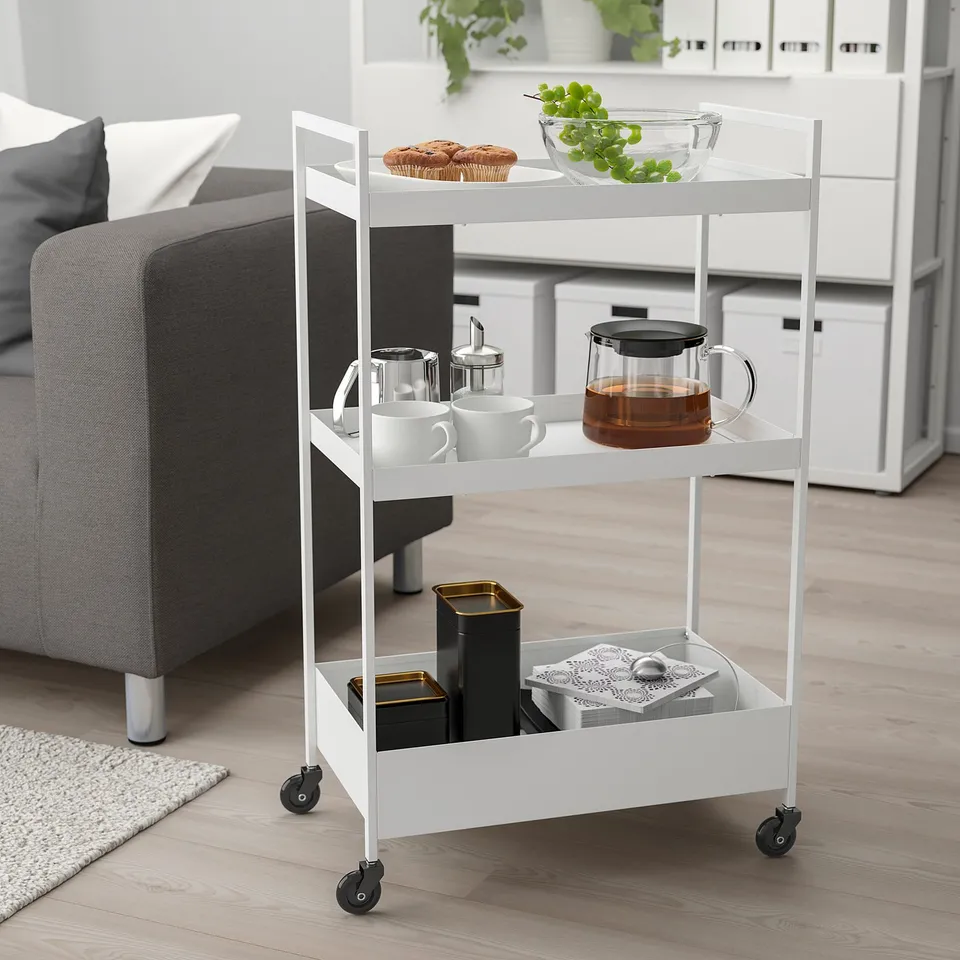 NISSAFORS white 3-shelf trolley with kitchen items, tea, snacks next to grey sofa.