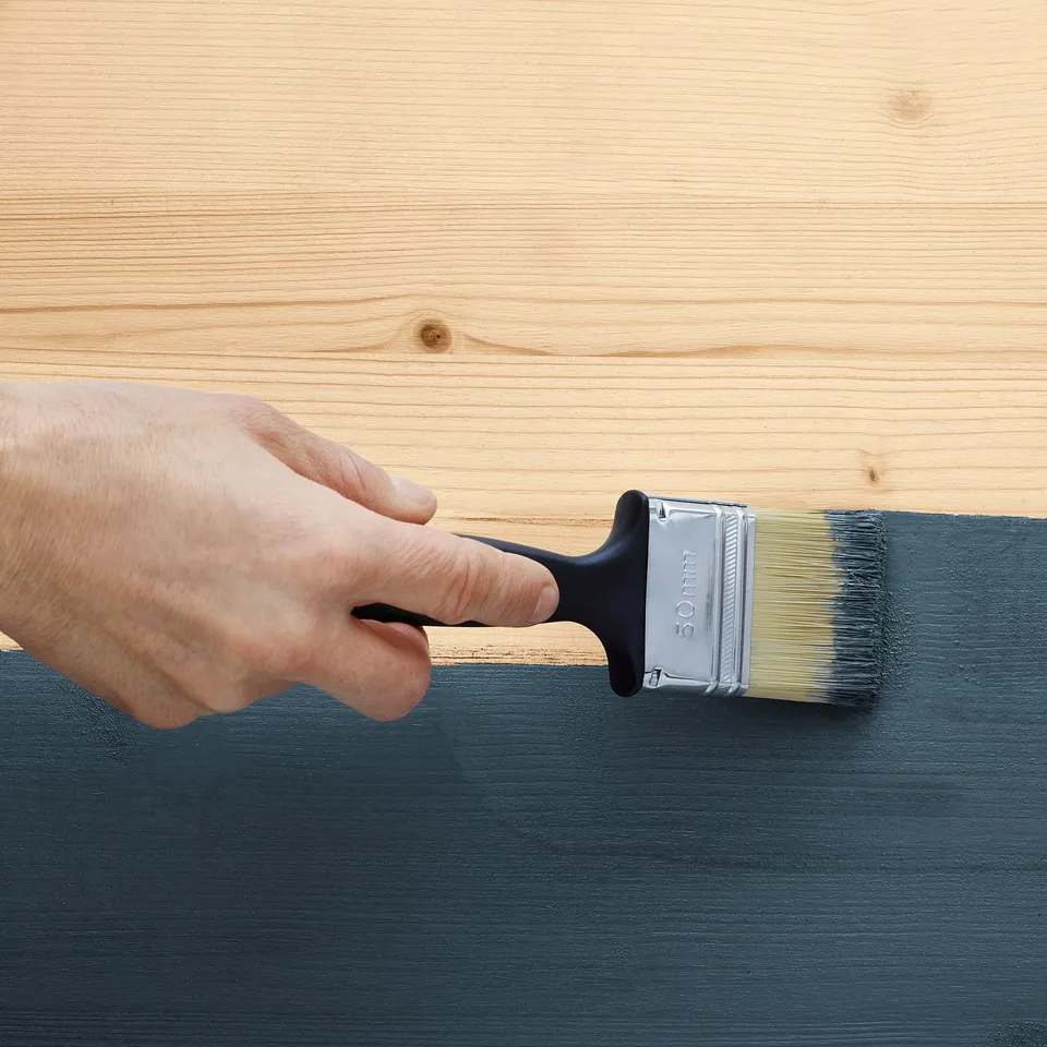 A hand uses a paintbrush to apply paint on wood, showcasing a diy project to finish an untreated wooden bed frame.