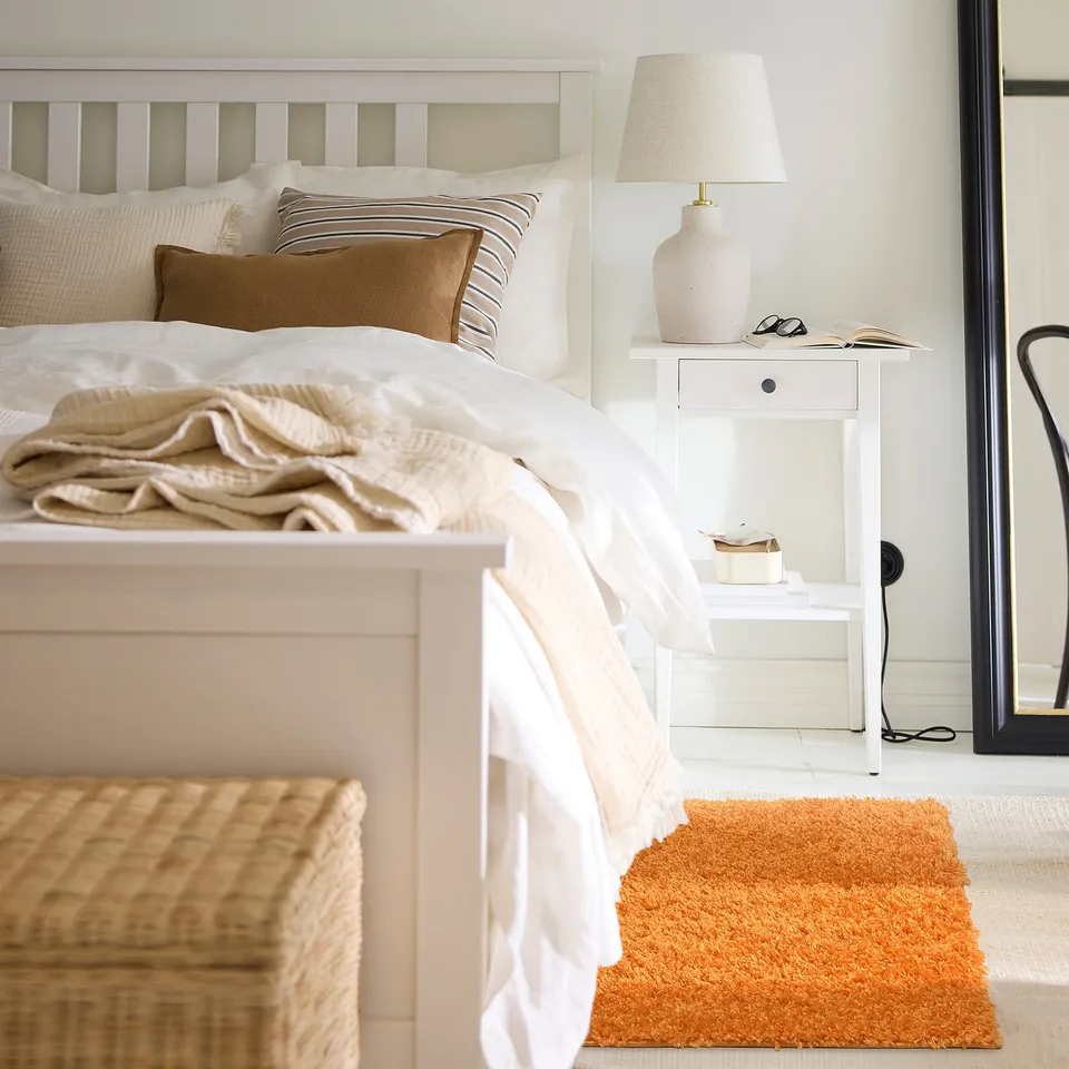 Bedroom with ÄRENDE orange rug, wicker chest, white bed, bedside table, lamp, and mirror; rug smaller than bed.