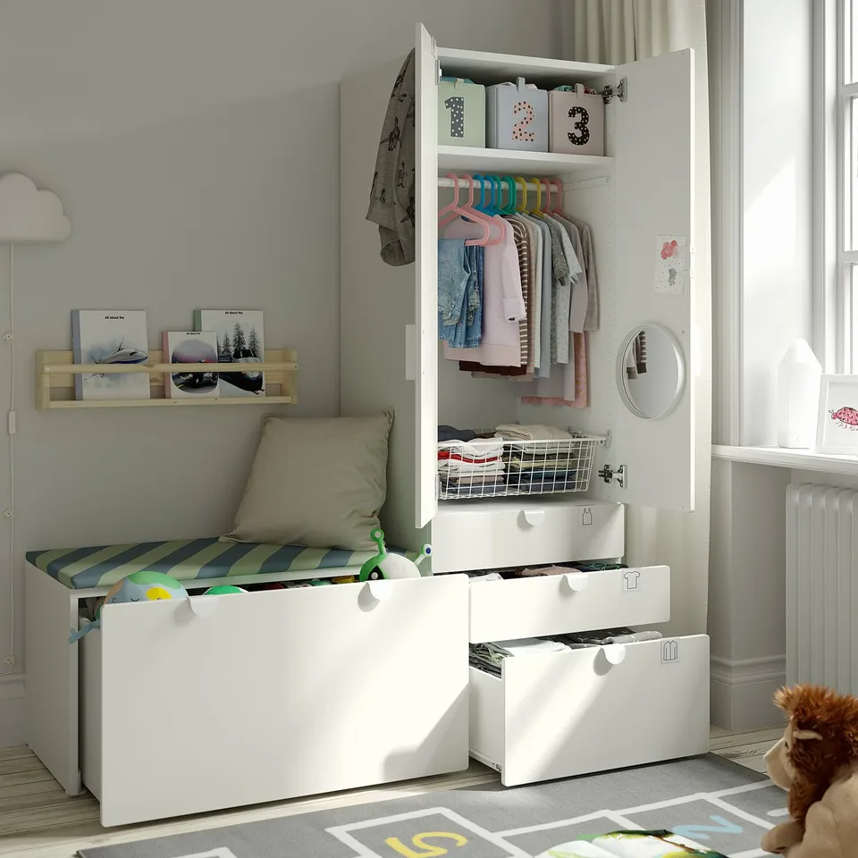 Cosy kids room with white storage unit: open wardrobe shows hangers, shelves, and drawers. Bench with cushions offers seating.