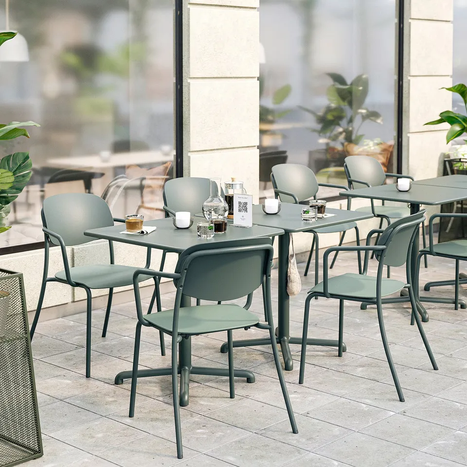 Modern outdoor cafe with green VISINGSÖ furniture. Tables have hooks, cups, and coffee sets. Simple, tough chairs. Tables are smaller than plants.