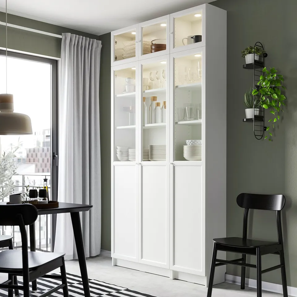White BILLY bookcase with glass doors dishes and decor. Tall, narrow, fits neatly by window, surrounded by dark chairs.