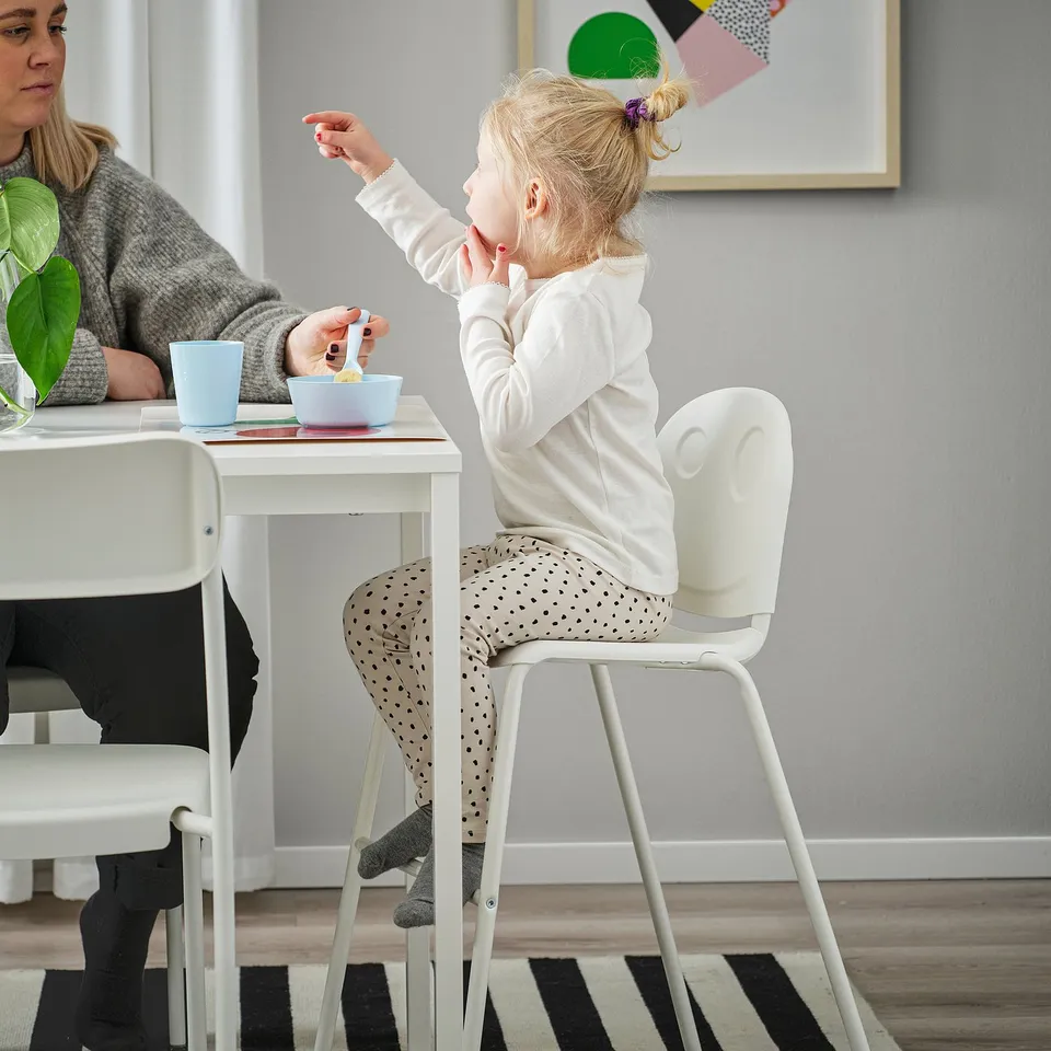 Child sitting on a VILDGÅS junior chair, eating from a bowl at a white table. Mum nearby, feet on chair.