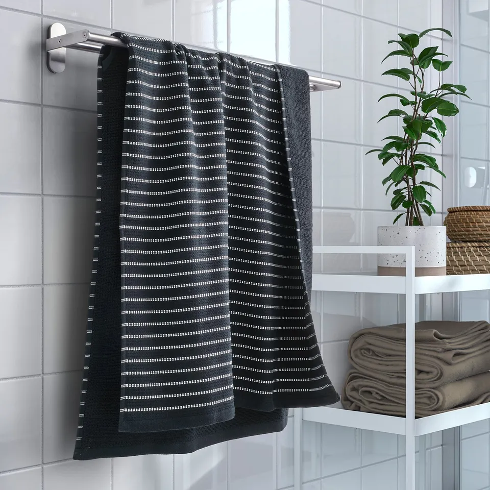 Black and white striped BROKGINST towel hangs near open window, surrounded by folded towels and a plant in a modern tiled bathroom.