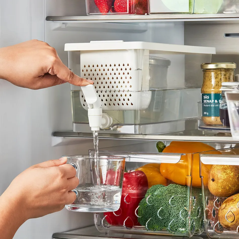 Person filling clear container with water from refrigerator dispenser, surrounded by vegetables and condiments.