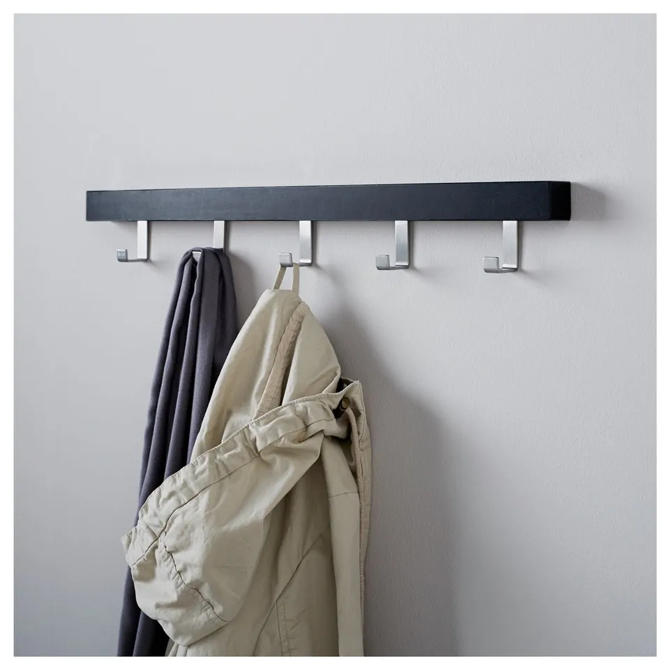 A black wooden coat rack with four metal hooks holds a grey and a beige jacket. The rack is mounted on a light-colored wall.