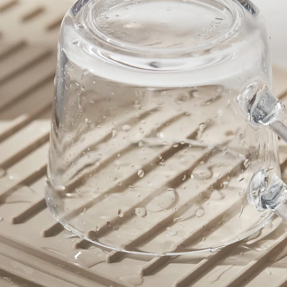 Clear glass with handles sits on a beige, ridged mat, likely a RINNIG drying mat, designed to catch water drips and keep dishes stable.