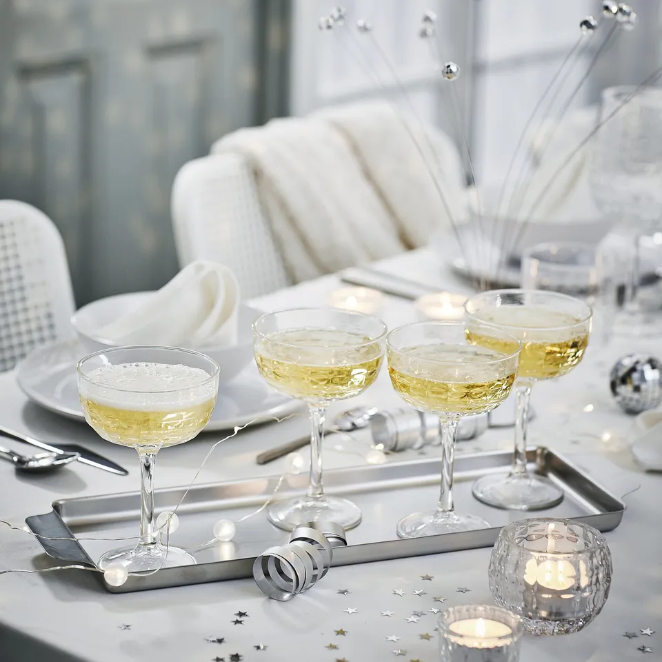 Elegant FRÖJDA champagne coupes, clear glass, wide bowls, on metallic tray with silver napkins, surrounded by festive decorations.