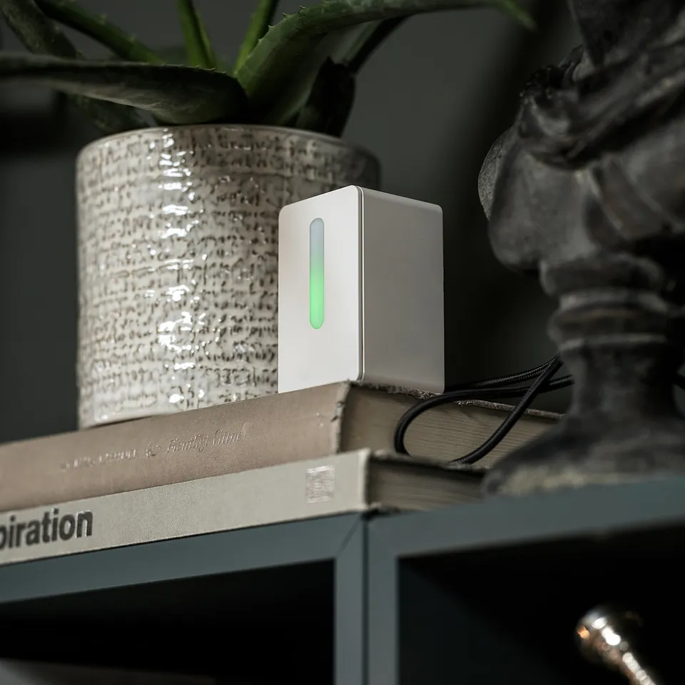 A small white sensor with a green light sits on books, next to a potted plant.