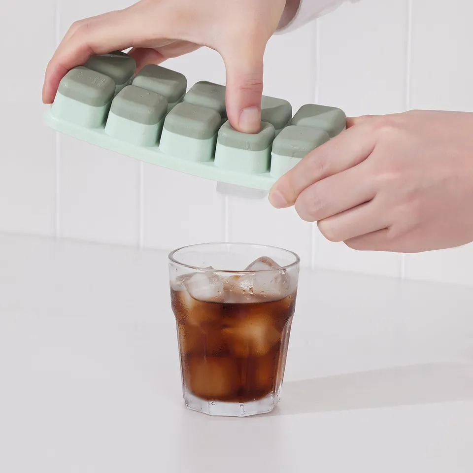 A person uses a spill-proof ice cube tray to fill a glass with ice cubes. The tray has a lid, preventing spills while transferring ice cubes.