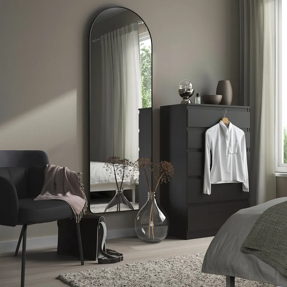 Modern bedroom with large, lean, black LINDBYN mirror, sleek dresser, cosy chair, and stylish decor.