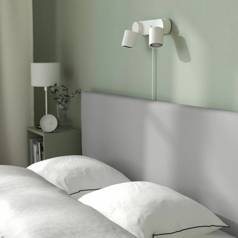 Clean, modern bed with storage. Wall light above, soft grey headboard. White pillows, green wall.