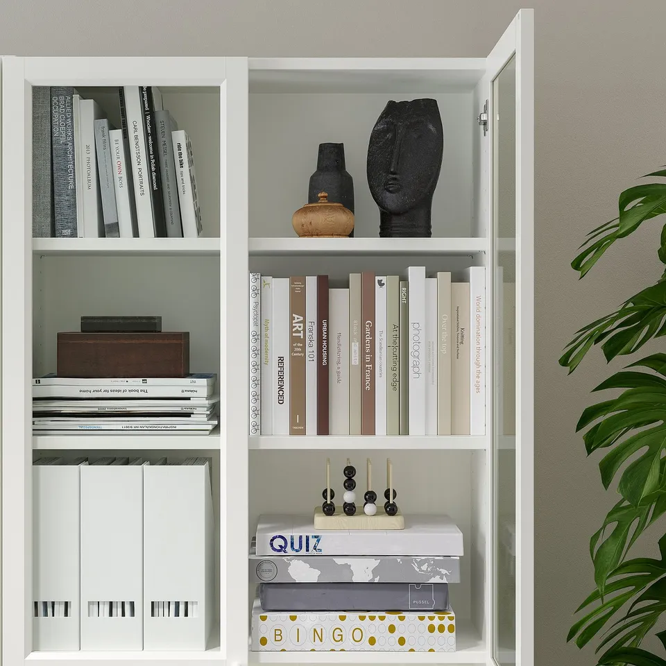 White BILLY bookcase with glass doors, displaying books, decorative items, and games, anchored to wall.