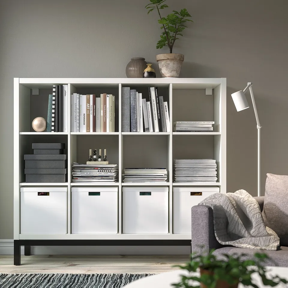 White KALLAX shelving unit with books, boxes, chair, lamp - grey wall.